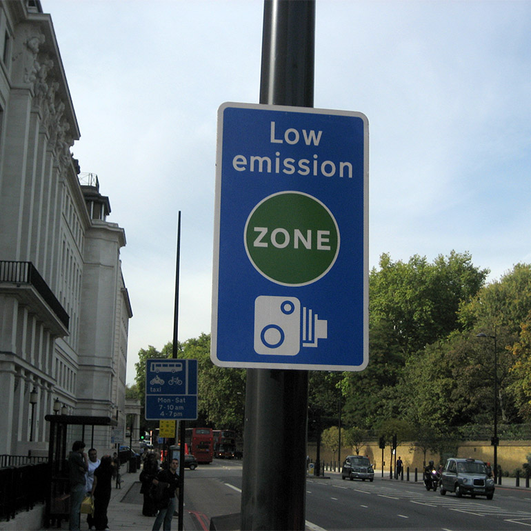 Photo of sign indicating a Low Emission Zone, enforced by camera along a city street. Taxies, buses and pedestirans use the street.