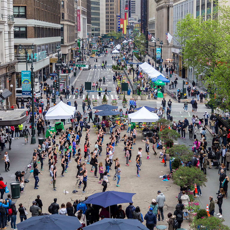 Image of a crowded pedestrian-access only street with booths, umbrellas, trees and what looks like a dance performance of two or three dozen participants taking place in the foreground. The downtown city street bends to the right in the background with more intersections, trees and popel walking throughout.