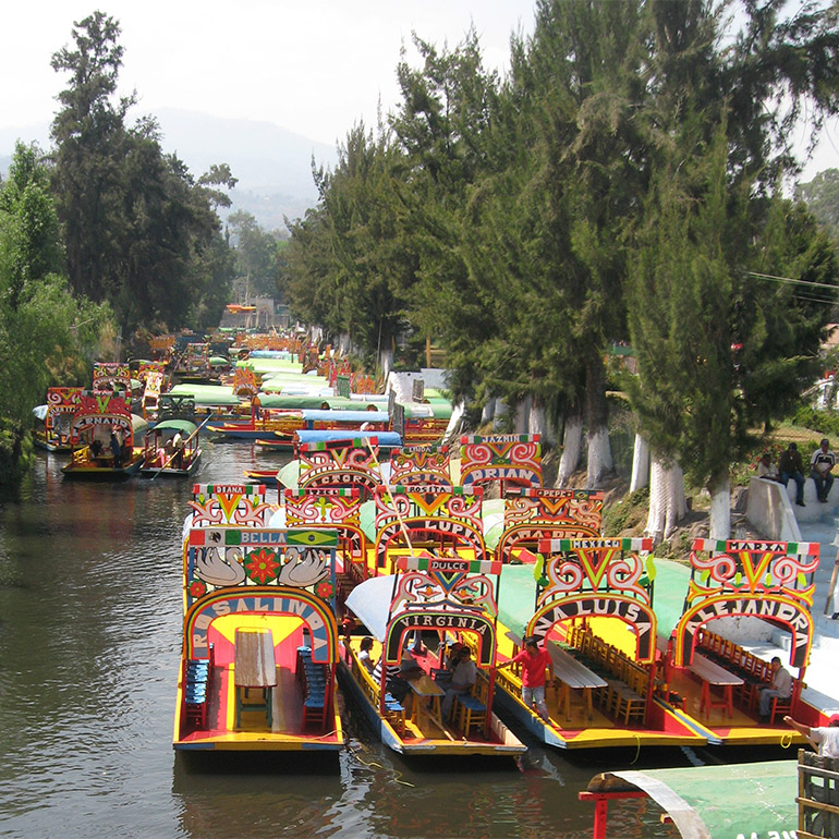 Dozens of small colorful boats with names such as Bella and Virginia travel along a river lined with large trees.