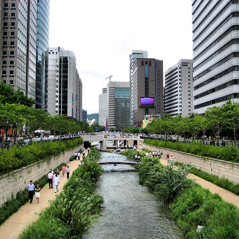 Photo showing a waterway passing through a canal with lush green foliage and pedestrian walkways on either side. Further along there are bridges of different sizes connecting the sides. In the background are half a dozen skyscrapers and what looks like an outdoor plaza with seating.