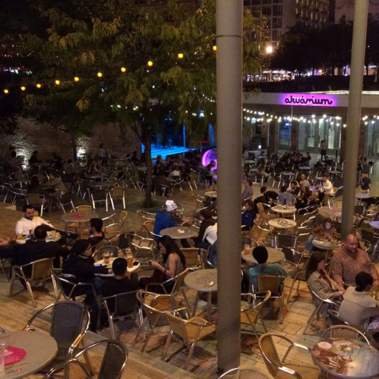 Aerial view image of an outdoor plaza with seating for nearby restaurants beneath trees and patio lights. Several dozen people sit in the space eating and drinking.