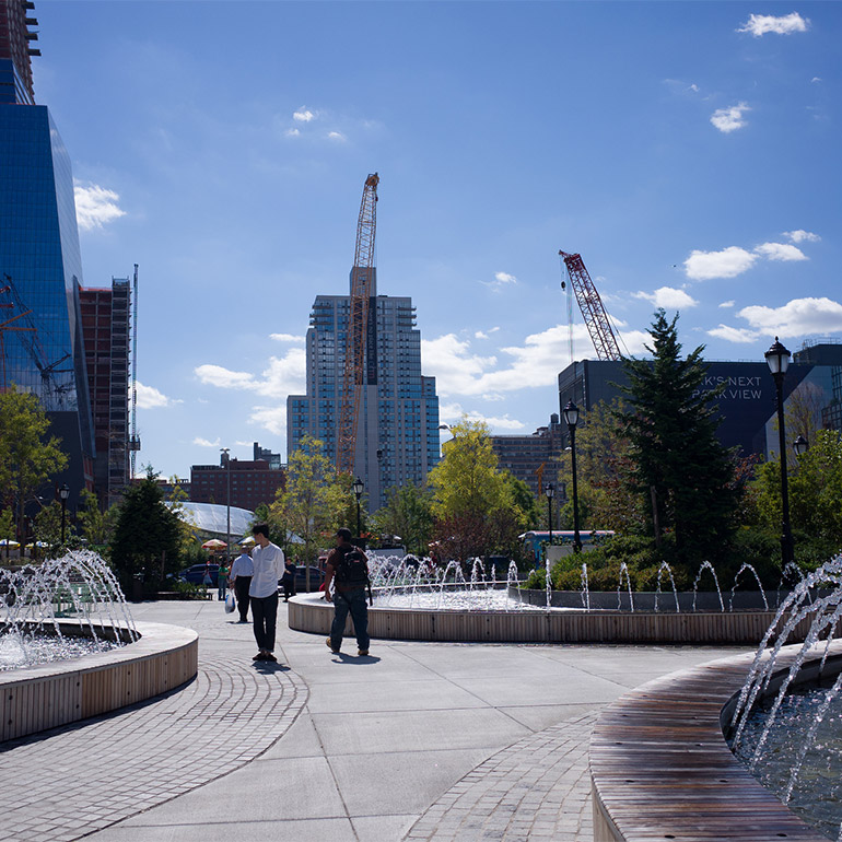 Image of an outdoor urban plaza with three water fountains and a walkway between them. Tall skyscrapers and cranes visible in the background. A few people are walking near the fountains in the center of the photo. There are trees on the outside of the plaza in the middle of the scene.