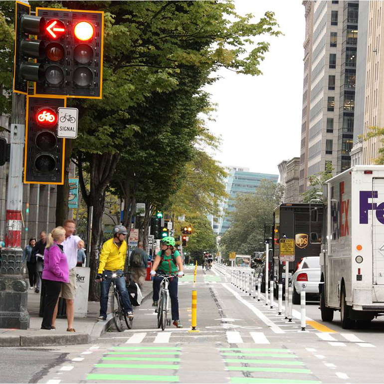 Image shows a street level view of a two way bike lane in the city, delivery trucks parked parallel to the lanes with a row of plastic bollards acting as a physical barrier. Two cyclists stopped at a red light are having a conversation and nearby them, two other pedestrians are waiting to cross the street. The street is lined by tall green trees on either side.