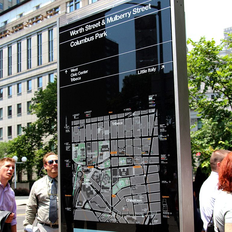Photo of a city map posted on the street with pedestrian wayfinding details. In the background a grey office building stands behind several trees.