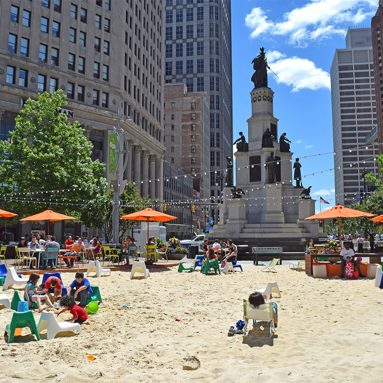 Image of a sandy park with chairs, umbrellas and tables in the middle of a city square. There is a large statue and tall, pale-colored office buildings in the background. Light green trees line the street on either side of the statue which is on the edge of the sandy park. People are seated at tables, under ubmrellas and children sit in and play with the sand.
