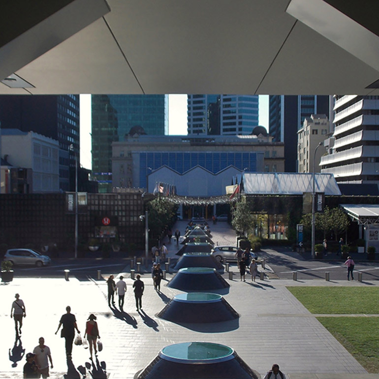 Photo of a public plaza in a downtown core with an overhang and pedestrians walking along a central walkway in the sun toward a street and larger building in the background. There is a corner of a lawn in the lower right hand corner of the photo.