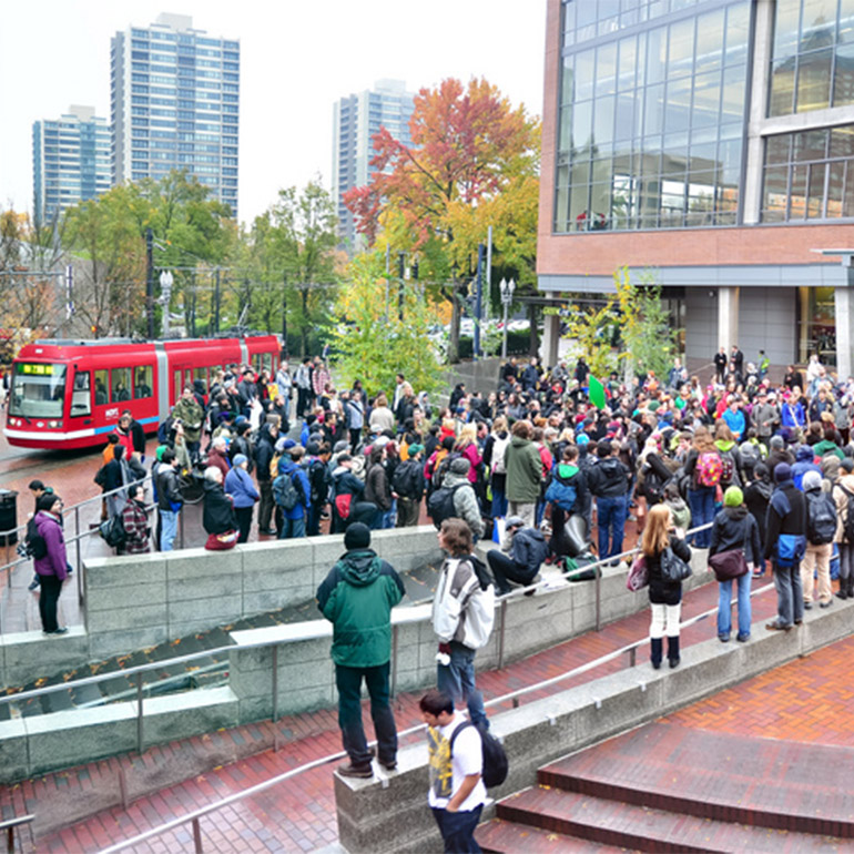 Image of a gathering of several dozen people in a downtown area plaza with a park and office buildings in the background. Between the group and the trees is a brigh red street car on its route.
