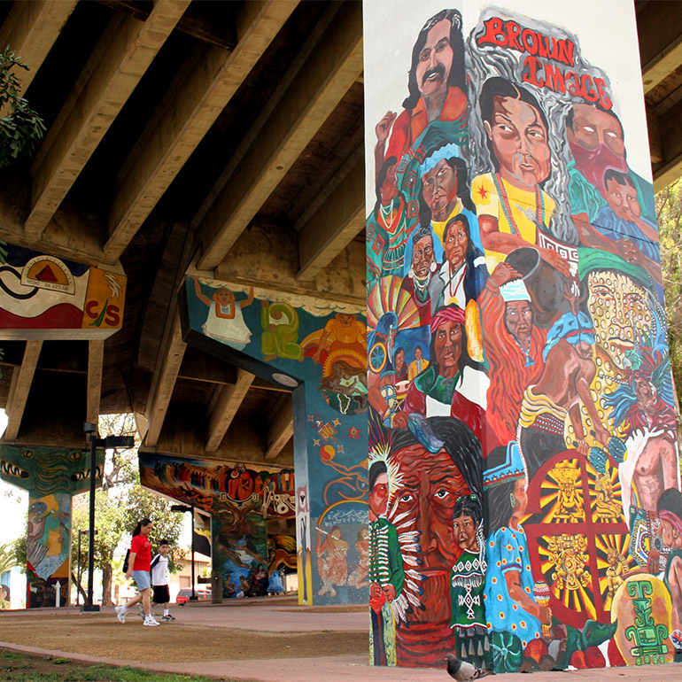 Image of children walking beneath a highway overpass, the pillars of which are covered in colorful murals depecting people of color and symbols from a diverse array of cultures.