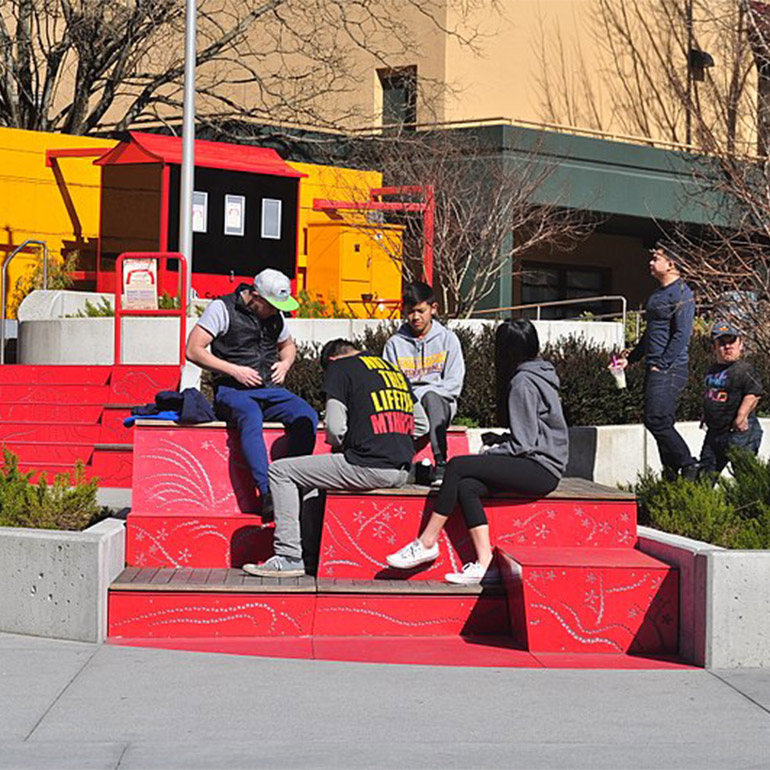Several people sit in the sun in a park with large red stairs as part of the landscaping. It is an open-air gathering space with trees and bushes in the background.