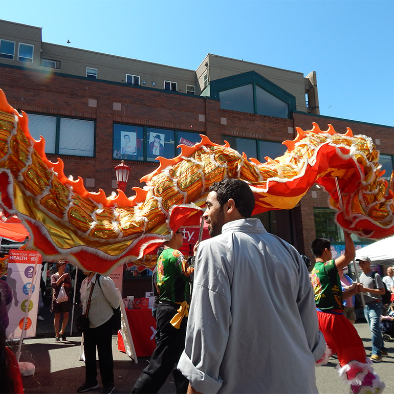 Photo shows a large cloth dragon puppet is paraded through the street at a festival in an urban setting. A person in the foreground observes the puppet's passing in front of them. There is a tall brick building in the background and blue skies above.