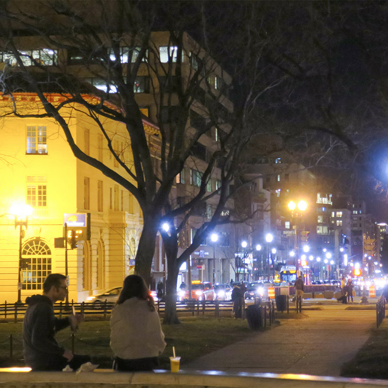 Image shows a nighttime view of a city street with street lamps on either side. A couple sits in a park beneath a large tree and face the street lamp-lined street.