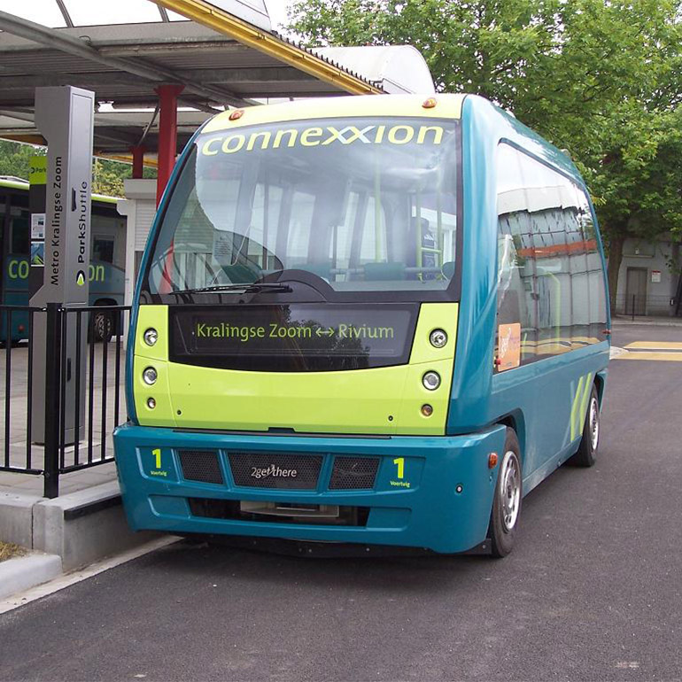 Image of a short, blue and green city bus with the word 'connection' spelled with two x's at the top of the windshield. It is parked at a bus stop and another similar looking vehicle is parked in the background.