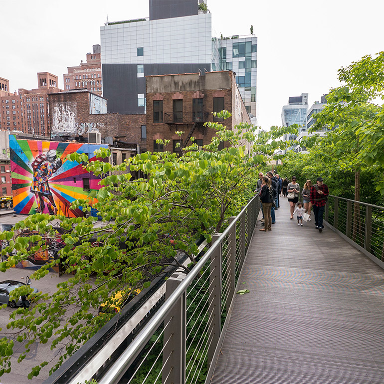 a group of people stand on the elevated highline pedestrian bridge in an urban setting surrounded by trees and with a streetart mural in the background