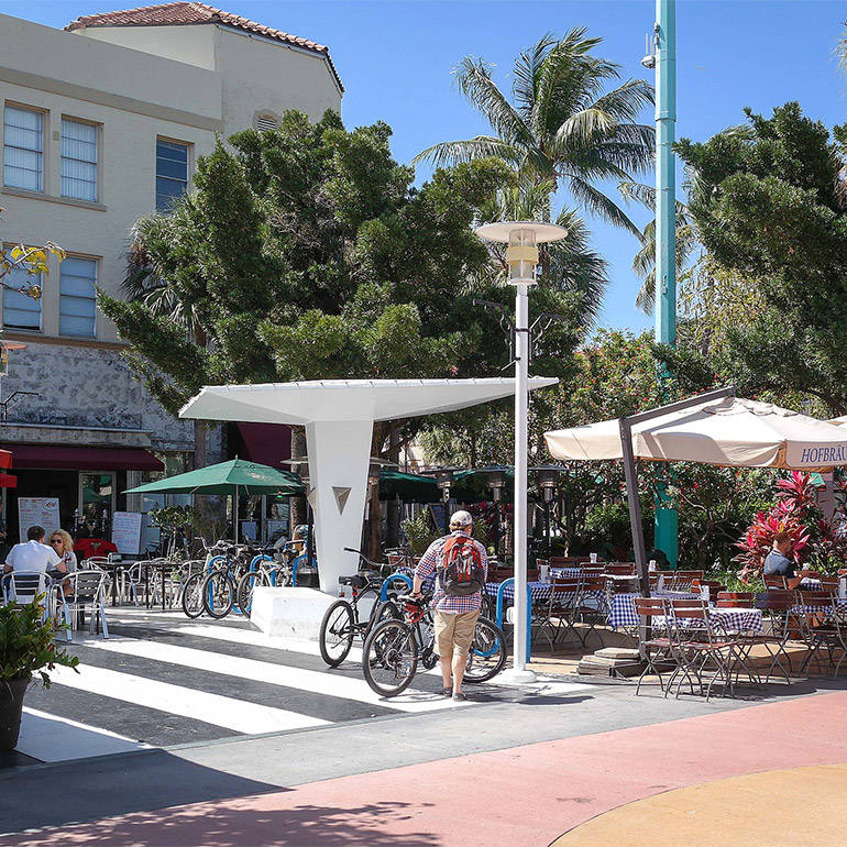 Person wearing shorts and a baseball hat walks bicycle to a bike rack in a sunny outdoor plaza with several tables and chairs with people dining outdoors on a sunny day. In the foreground, the ground is painted different colors, indicating a motorized-vehicle zone. In the background, tall trees including palm trees create shade over some of the seating area.