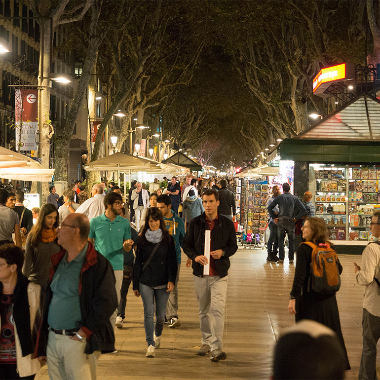 Photo of a crowded pedestrian-only street with shops, umbrellas extending into the background. Tree-lined walkway is lit up at night by streetlight and lights from the shops.