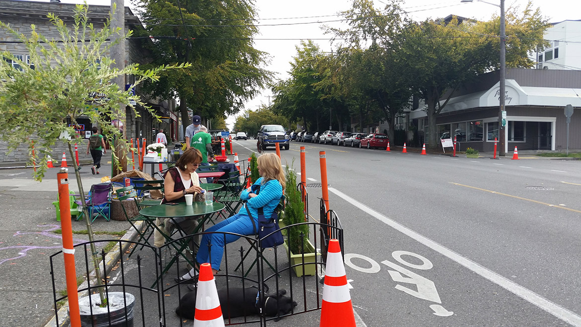 Two people sit at a table in a pop-up park next to a street with a protected bike lane