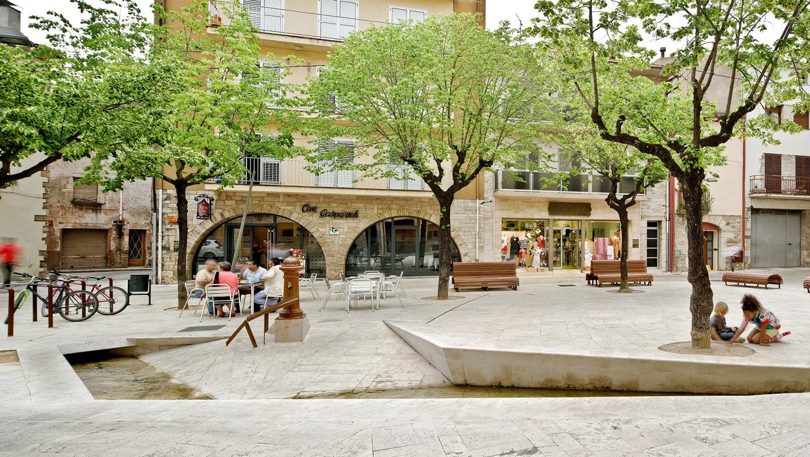 People sitting at tables in an outdoor plaza beneath green trees. Benches are also available for sitting around a central area.
